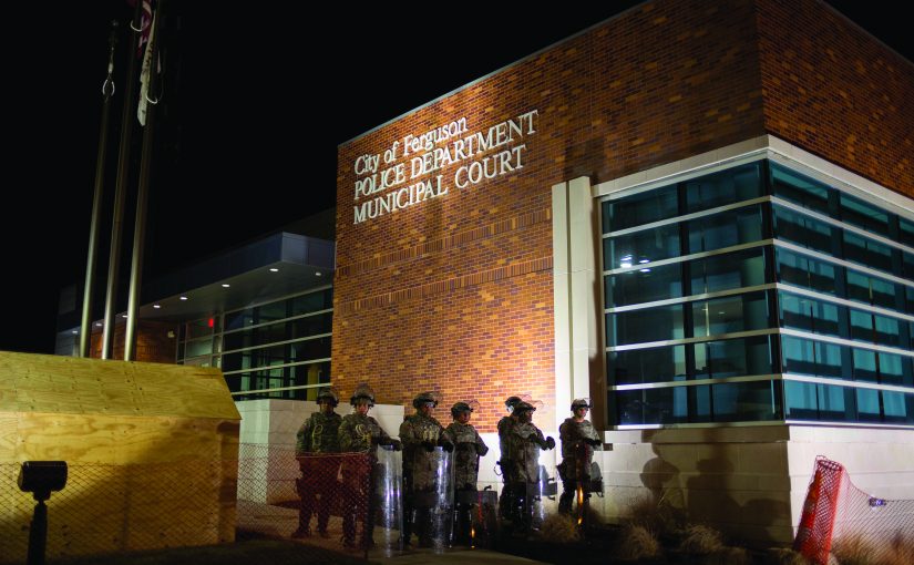 Seven uniformed officers with shields stand at night outside City of Ferguson Police Department and Municipal Court; flags and wooden structure nearby