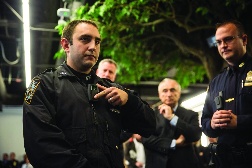 Law enforcement officer demonstrates body camera to colleagues indoors under artificial lighting; tree and equipment visible in background.