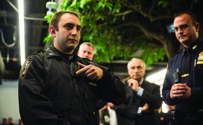 Law enforcement officer demonstrates body camera to colleagues indoors under artificial lighting; tree and equipment visible in background.