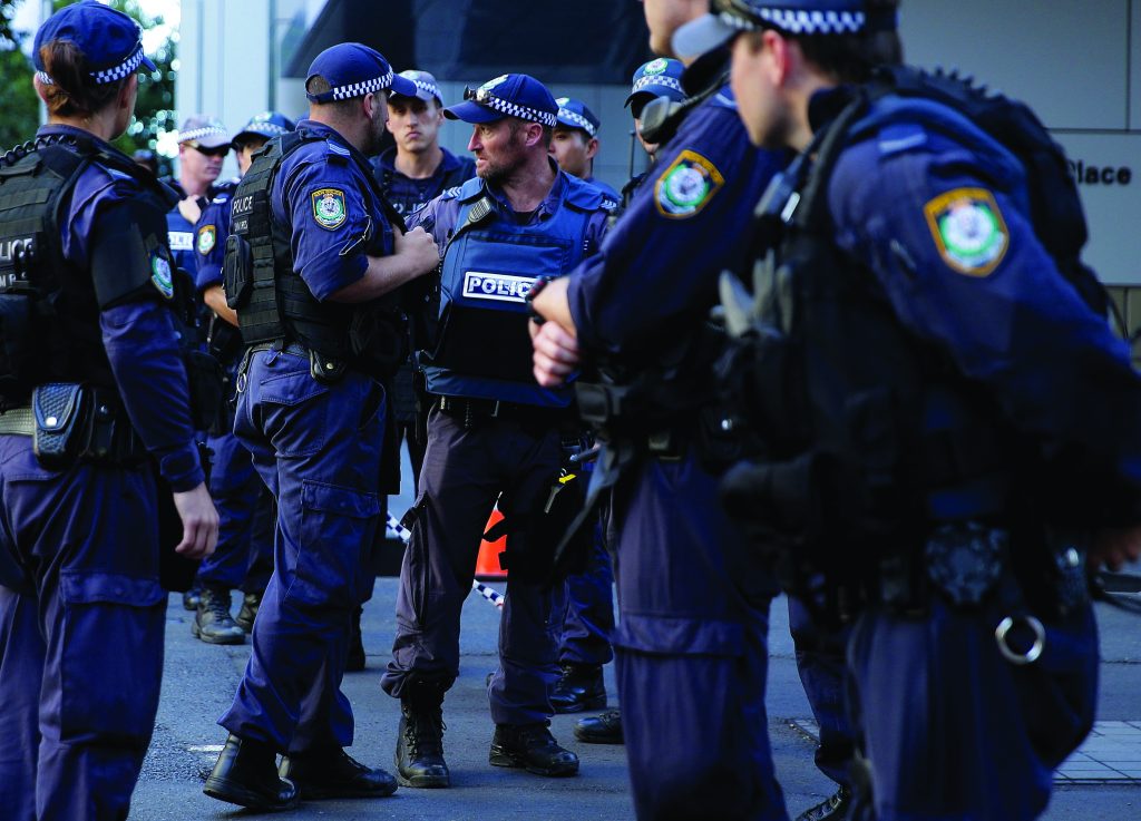 Multiple uniformed police officers wearing dark tactical vests, radios, duty belts, and checkered caps stand in a street briefing formation, with agency patches, body armor, and communication gear visible outside a commercial building