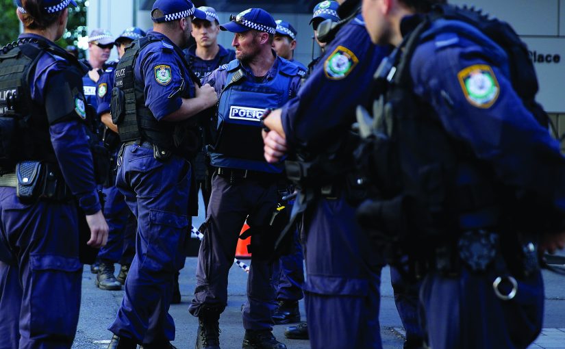 Multiple uniformed police officers wearing dark tactical vests, radios, duty belts, and checkered caps stand in a street briefing formation, with agency patches, body armor, and communication gear visible outside a commercial building