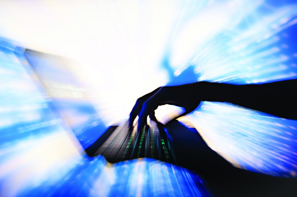 Silhouetted hands type on a laptop as bright light shines through the screen, with blue motion streaks forming a dynamic digital effect around it