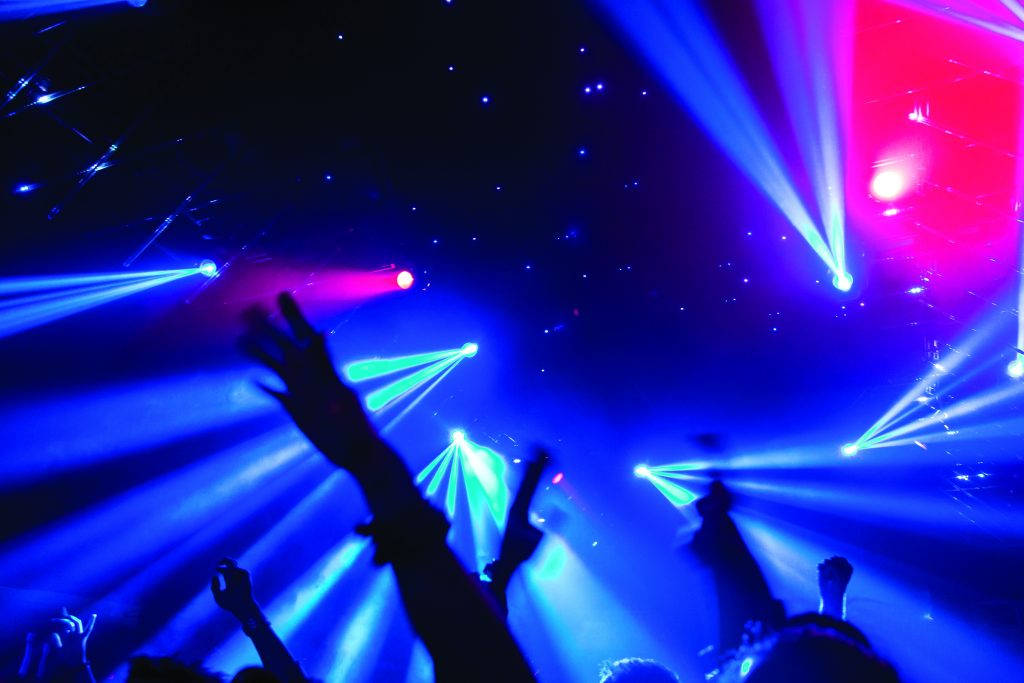 Concert scene with blue and pink spotlights on stage, silhouetted raised hands in foreground, and starry backdrop creating a vibrant, energetic setting