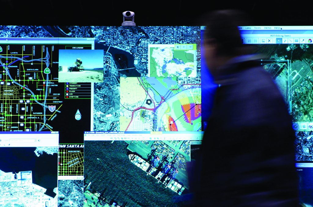 Large digital wall displays layered maps, satellite imagery, data panels, and street grids as a person stands nearby reviewing real‑time geographic information systems