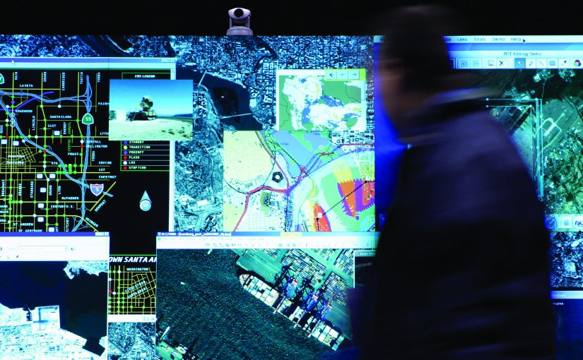 Large digital wall displays layered maps, satellite imagery, data panels, and street grids as a person stands nearby reviewing real‑time geographic information systems