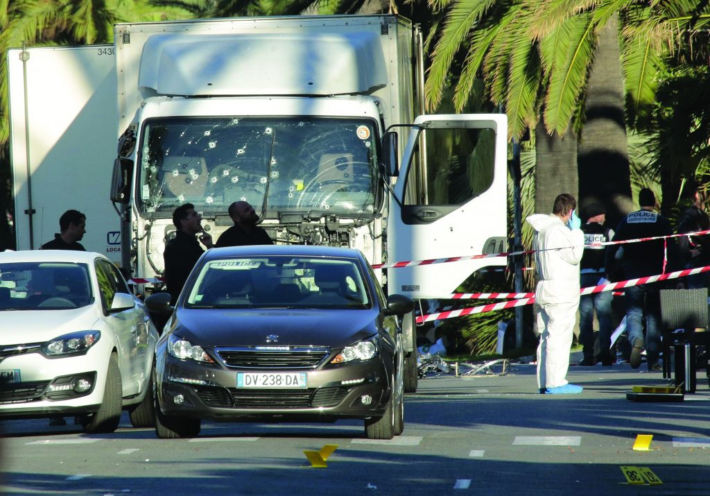 Crime scene with bullet-riddled white truck, police tape, forensic and law enforcement personnel; two cars in front, palm trees in background.