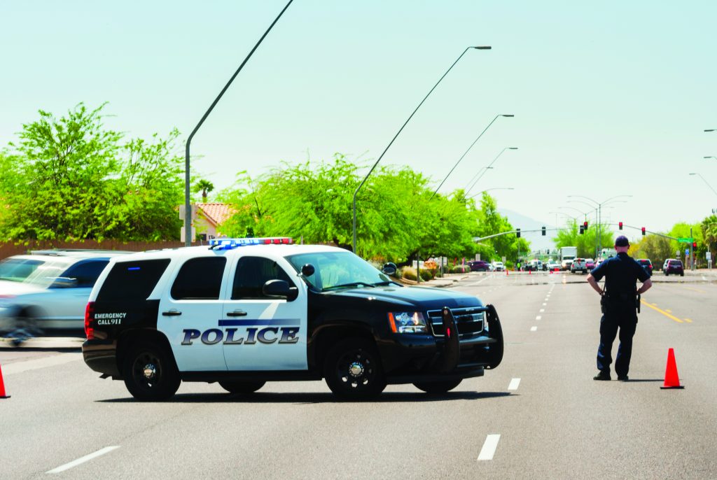 Marked black-and-white police SUV blocks a multi-lane roadway during a traffic closure, with emergency lights activated, traffic cones, streetlights, trees, distant signals, and an officer standing near the center of the road
