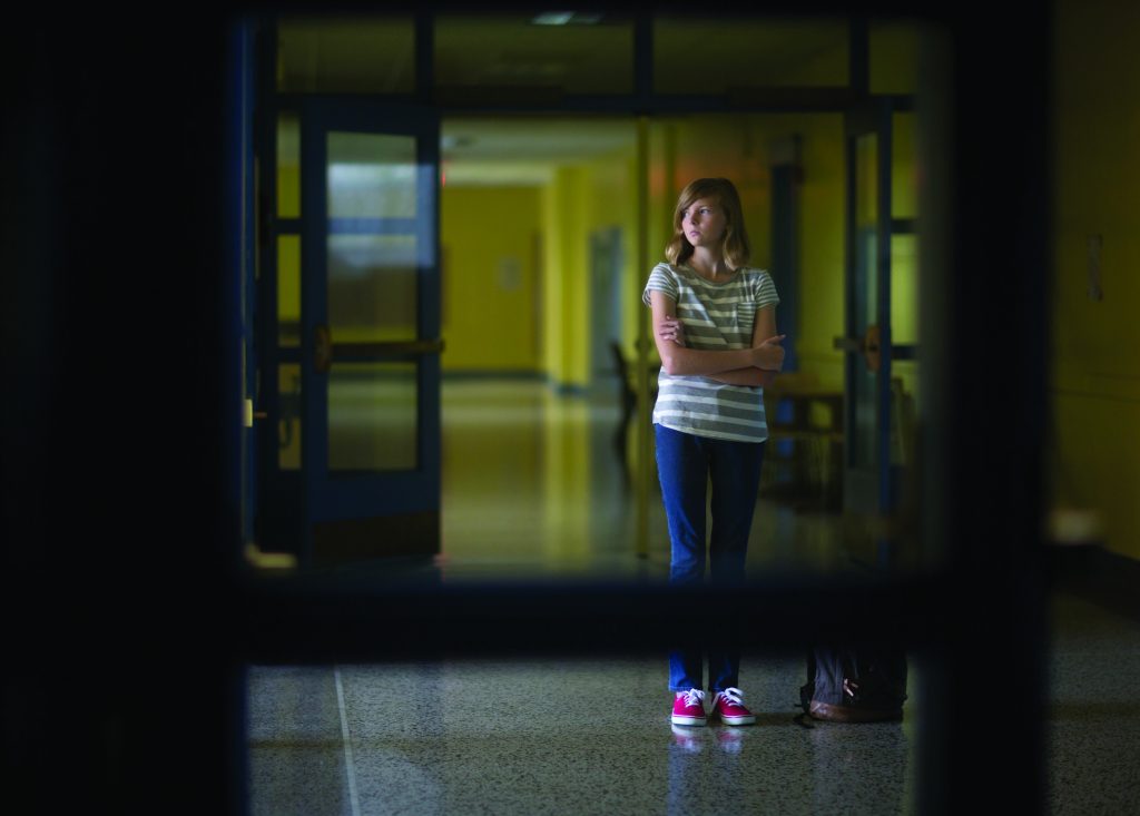 Individual stands in dim hallway with yellow walls, blue double doors, striped shirt, jeans, red shoes; arms crossed, near wall-mounted equipment.