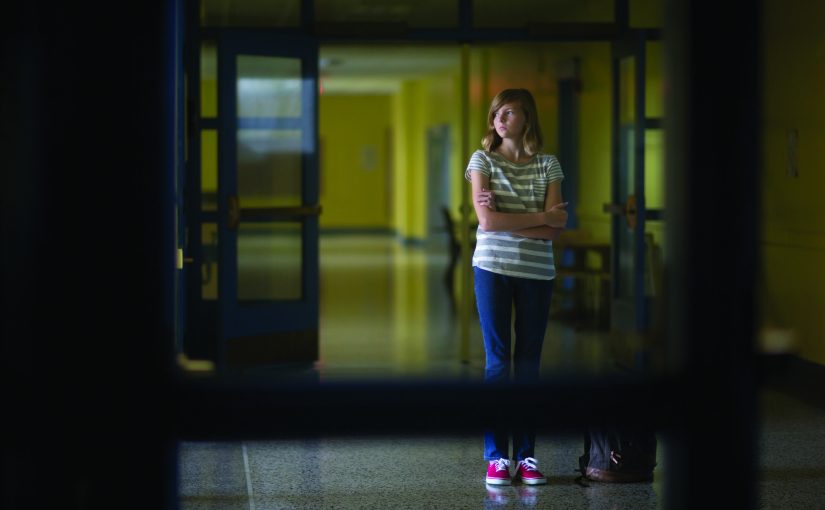 Individual stands in dim hallway with yellow walls, blue double doors, striped shirt, jeans, red shoes; arms crossed, near wall-mounted equipment.