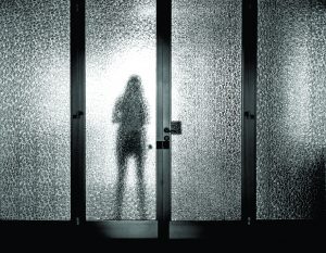 Silhouette of person behind textured glass doors; black and white image with blurred, high-contrast effect creating a mysterious atmosphere