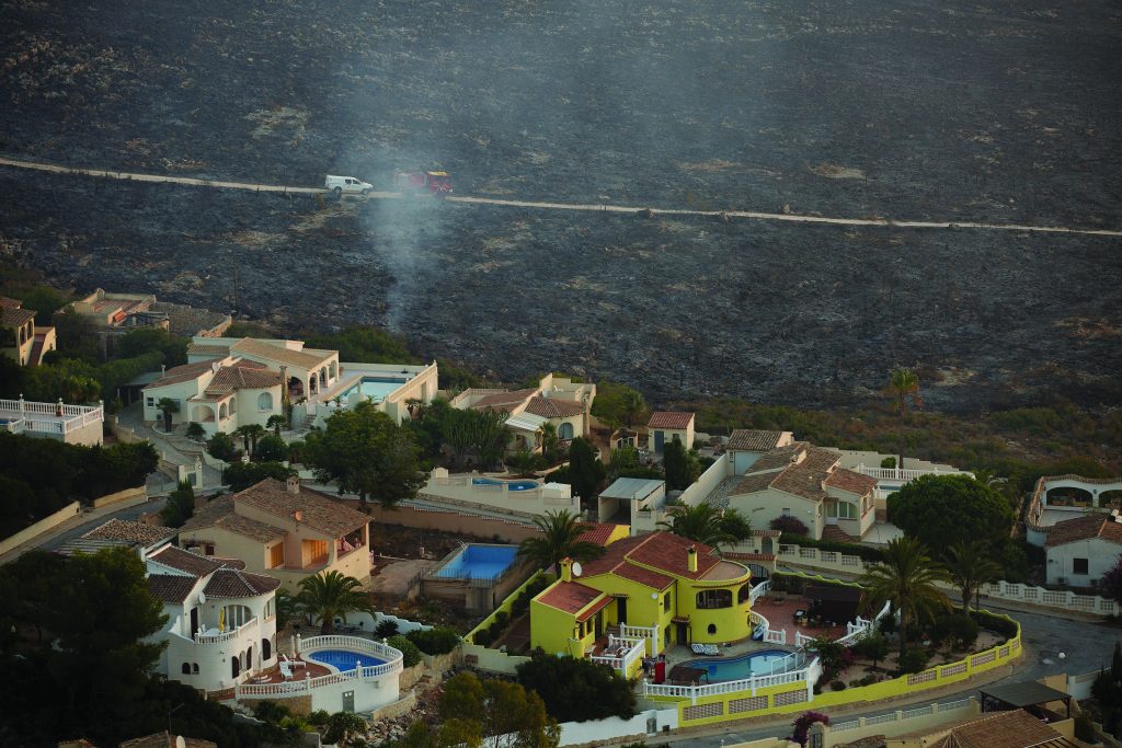 Row of Mediterranean-style homes with pools and palm trees near a hillside; smoke rises in background where emergency vehicles are parked on a dirt road.