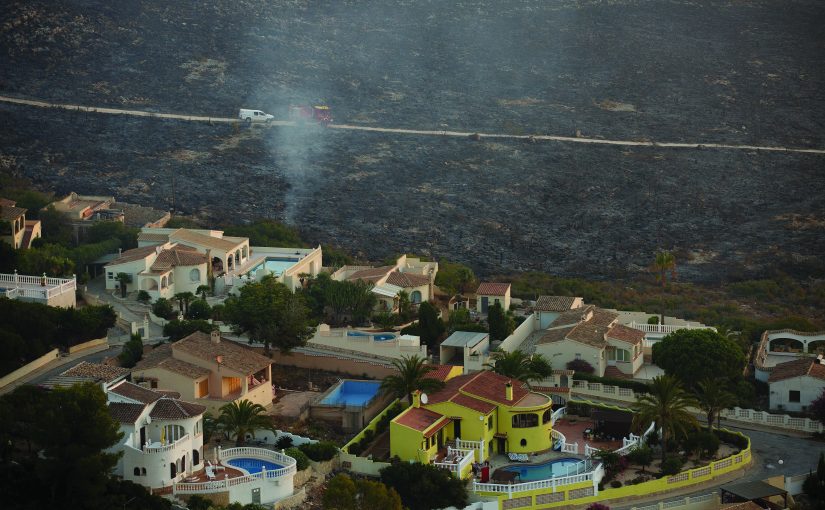 Row of Mediterranean-style homes with pools and palm trees near a hillside; smoke rises in background where emergency vehicles are parked on a dirt road.