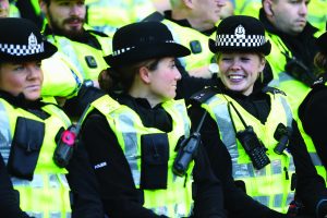 Uniformed police officers wearing high‑visibility vests and black helmets stand closely together in formation, with radios, body cameras, insignia, and winter gear visible
