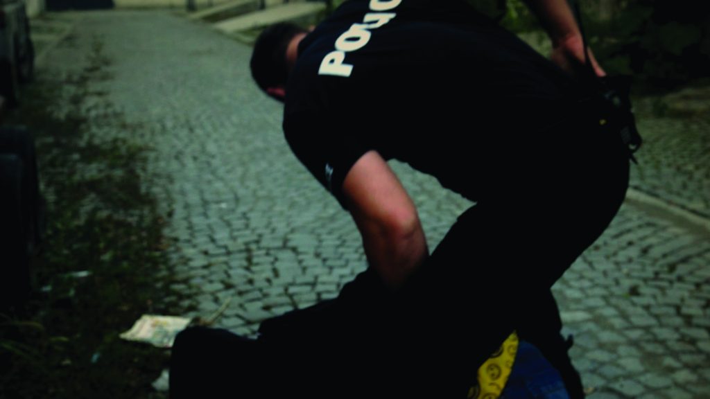 Officer in uniform restrains individual on cobblestone street, kneeling on back, securing arm; scene shows tactical control in public setting.