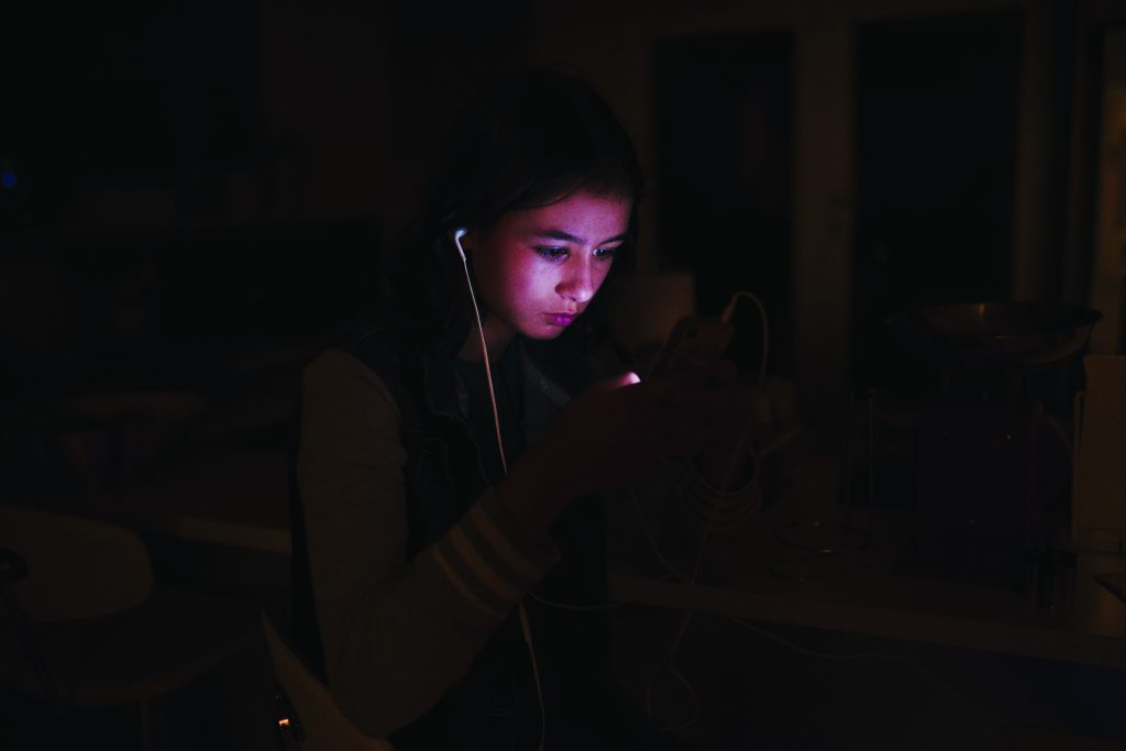 Person sitting indoors holding a smartphone with earphones plugged in, screen glowing in dim light, wires visible, background showing kitchen counter