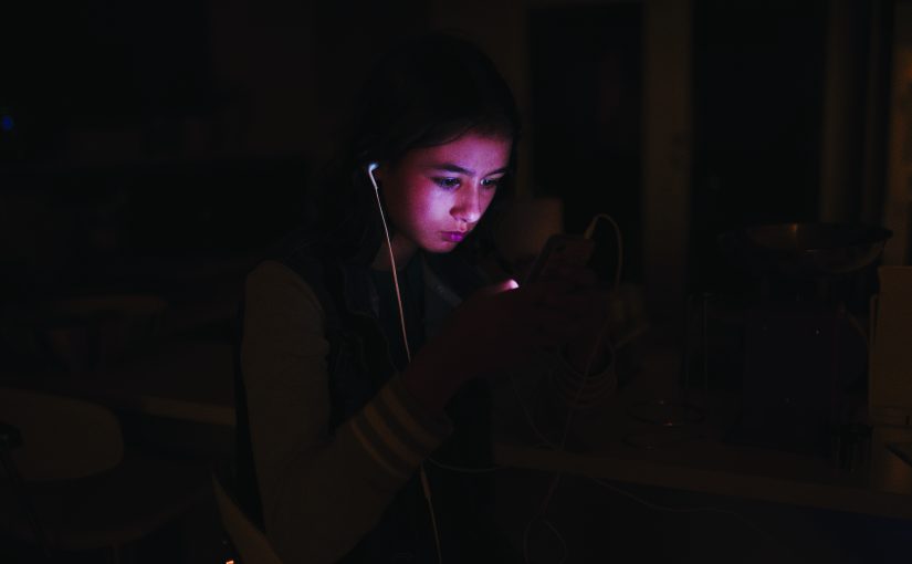 Person sitting indoors holding a smartphone with earphones plugged in, screen glowing in dim light, wires visible, background showing kitchen counter