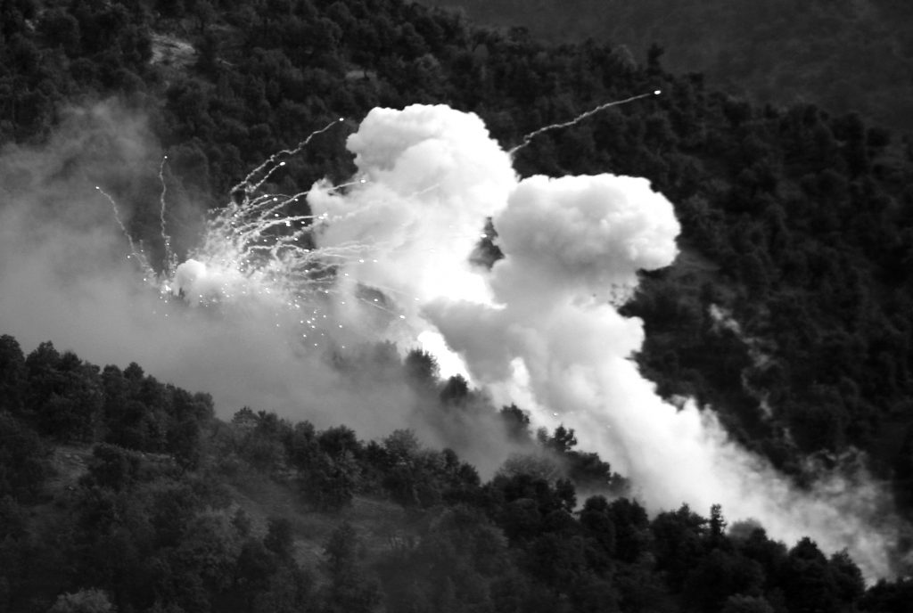 Large plumes of white smoke rise above a forested hillside as bright sparks and streaks arc outward, obscuring parts of the trees in a mountainous landscape