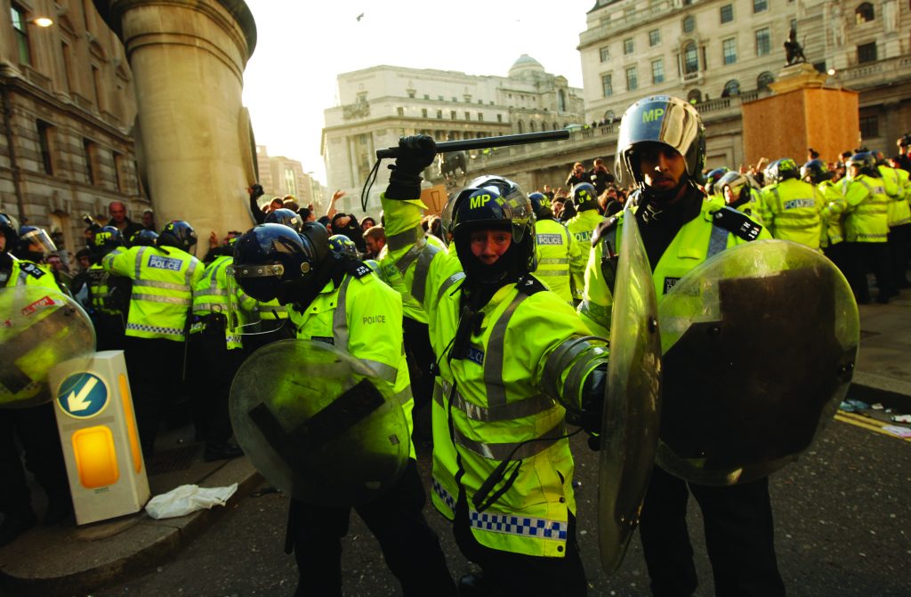 Police in high-visibility jackets, helmets, and shields form a line during urban protest; buildings and additional officers visible in background