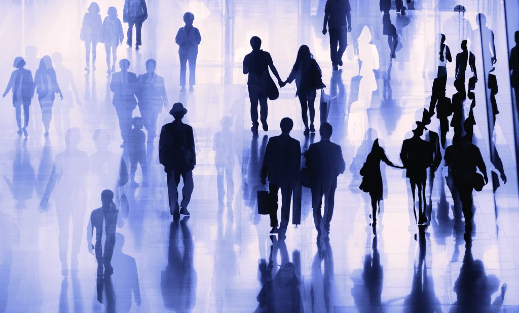 Silhouetted figures walk on reflective surface, casting shadows; some carry bags or hold hands; soft backlight emphasizes movement and direction.