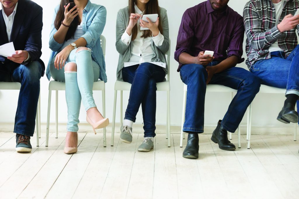 Diverse black and white people sitting in row using smartphones tablets, multiracial men and women waiting for job interview, human resources, employment or customers and electronic devices concept