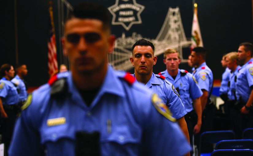 Uniformed officers in light blue shirts gather inside an event hall, with shoulder patches, radios, red epaulets, flags, chairs, and a bridge mural