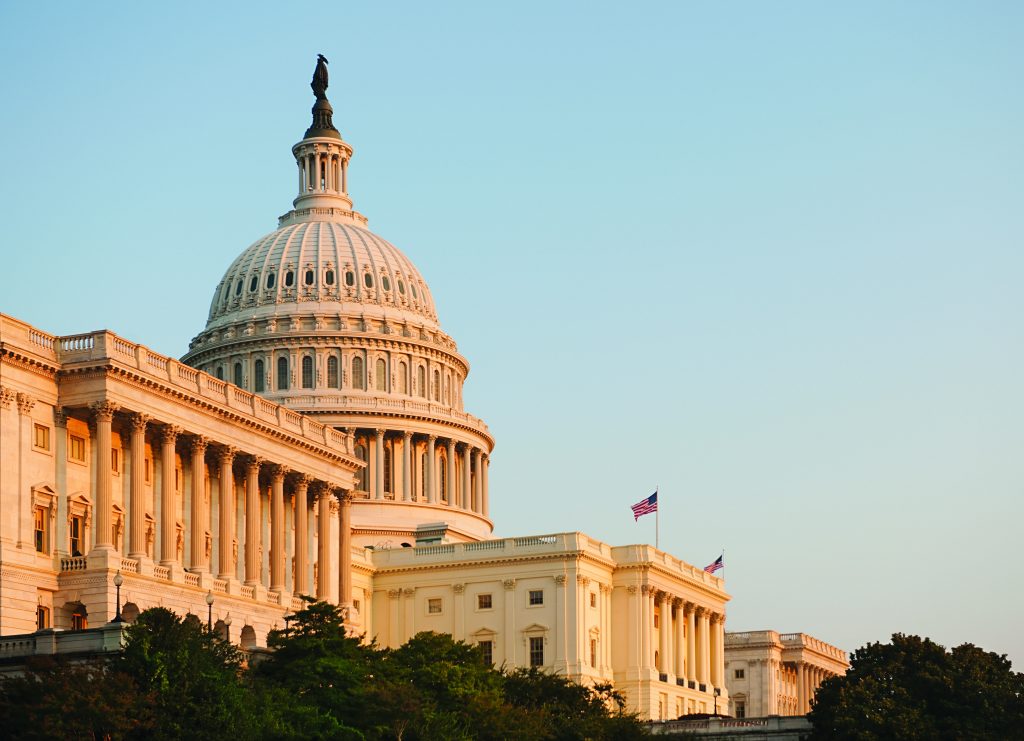 U.S. Capitol building with iconic dome and adjacent structures, lit by warm sunlight under clear sky; architectural details clearly visible