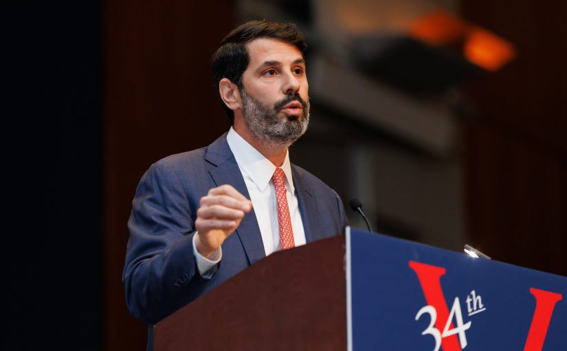 Speaker in suit stands at a podium with microphone, gesturing while delivering remarks at a formal conference stage, with large signage, lighting, and auditorium backdrop visible