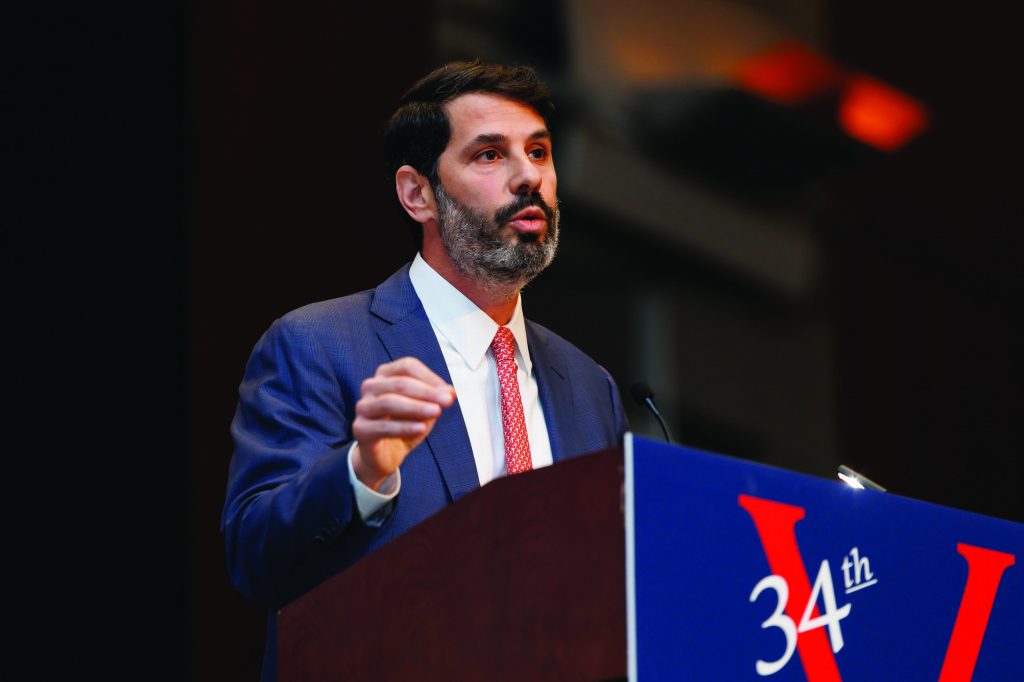 A suited speaker stands at a podium with a microphone, raising one hand while addressing an audience, with a large sign showing ‘34th’ and stage lighting visible behind