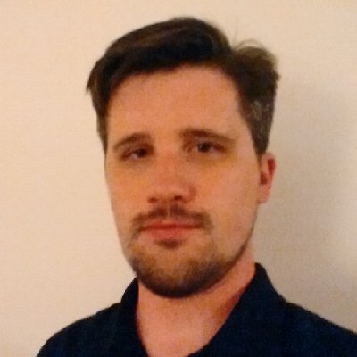 Individual with short, styled hair and trimmed beard wears dark collared shirt; posed against plain beige background for anonymity.