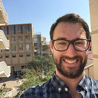 Person wearing a blue and gray checkered button-up shirt standing outdoors with beige brick buildings, balconies, and clear blue sky in the background