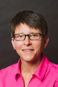 Person wearing a bright pink collared shirt with open neckline, short dark hair, and small stud earrings against a plain dark background