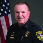 Uniformed officer stands before U.S. flag; visible badges and left-sleeve patch; dark background emphasizes patriotic and professional setting