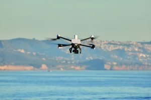 Quad‑rotor drone with camera gimbal hovers above calm water, its spinning propellers blurred, with distant coastal hillside, buildings, and hazy sky