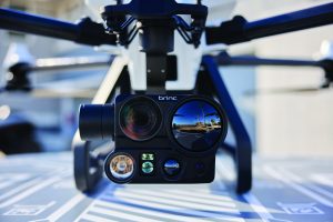 Close view of a drone gimbal shows dual camera lenses, flashlight, sensors, and Brinc branding mounted beneath quad arms with landing pad beneath
