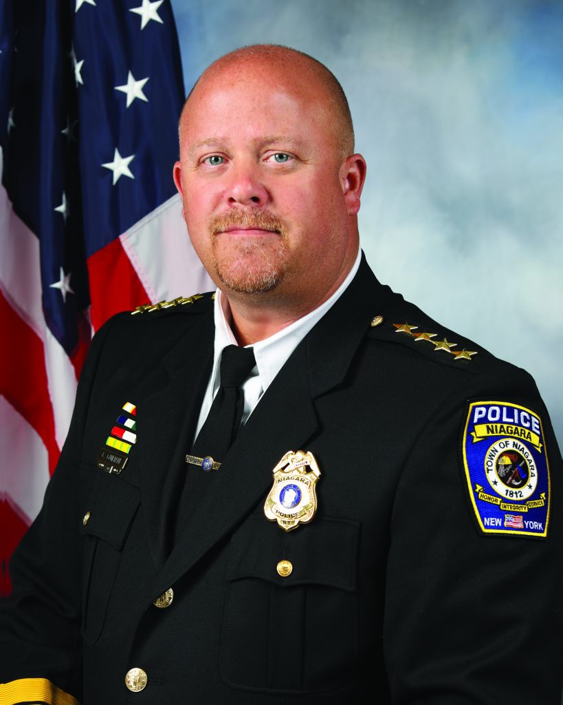 man in a police chief uniform is standing in front of an American flag. The man's face is blurred for privacy. He is wearing a dark navy blue police uniform with gold buttons and insignia, including a badge on the left side of his chest and several service ribbons above it. On his right shoulder, there are four gold stars indicating his rank. The patch on his left arm reads "POLICE NIAGARA FALLS NEW YORK" with an emblem and the year "1892". The background consists of a gradient from light to dark blue.
