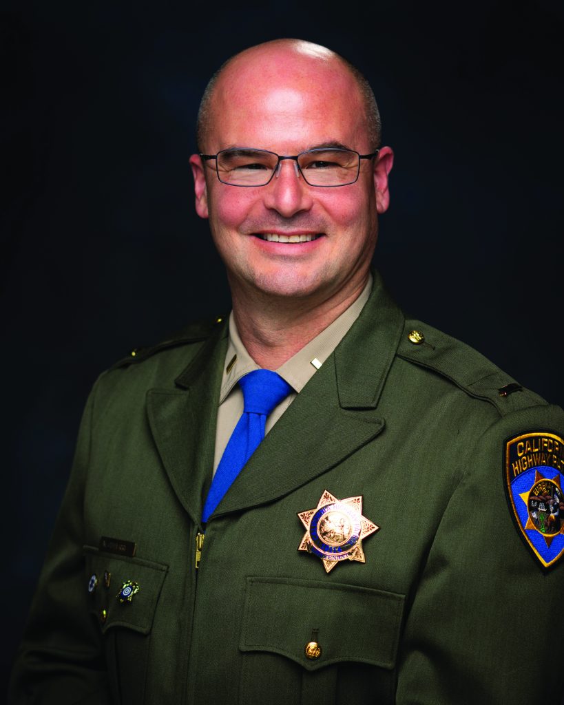 Studio portrait shows a uniformed law enforcement official wearing an olive dress jacket with badge, shoulder patch, blue tie, pins, and nameplate against a dark background