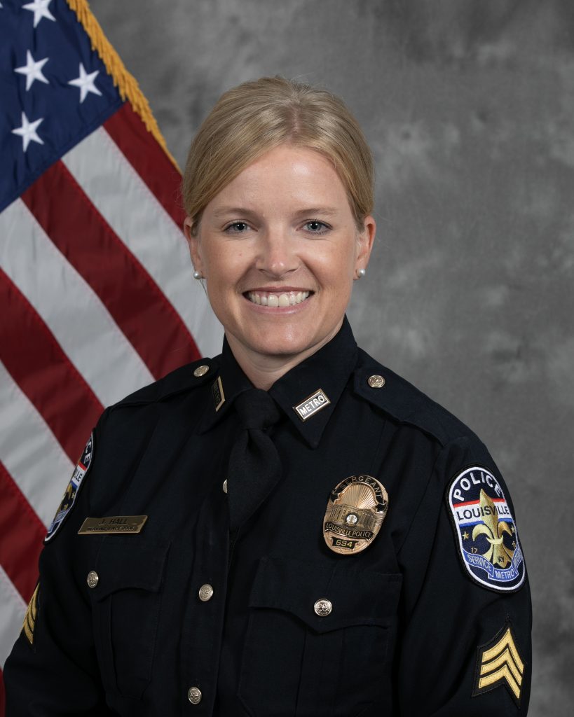 Police sergeant in formal dark uniform with badge, nameplate, and shoulder chevrons stands before an American flag backdrop and gray textured background
