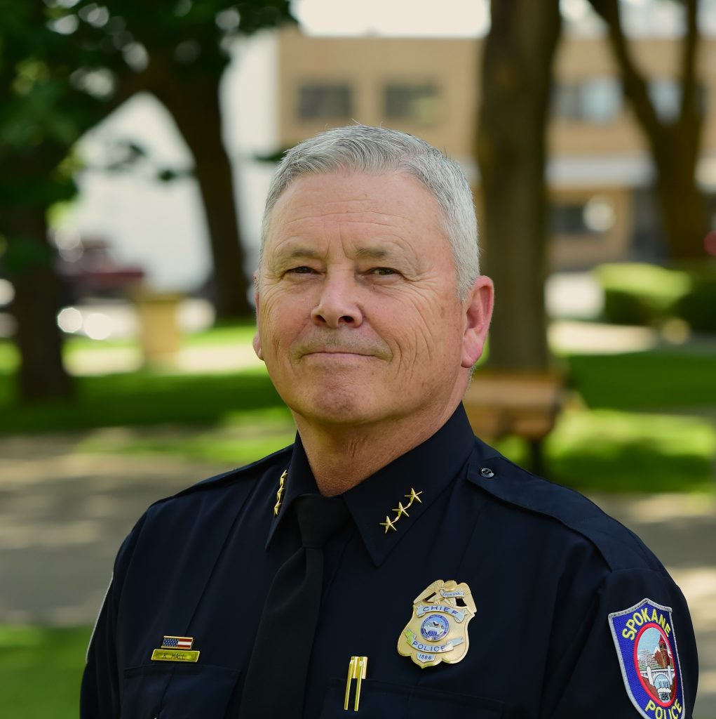 Uniformed police chief in black dress uniform with gold collar stars, Spokane Police badge, nameplate, ribbon bar, and shoulder patch stands outdoors.