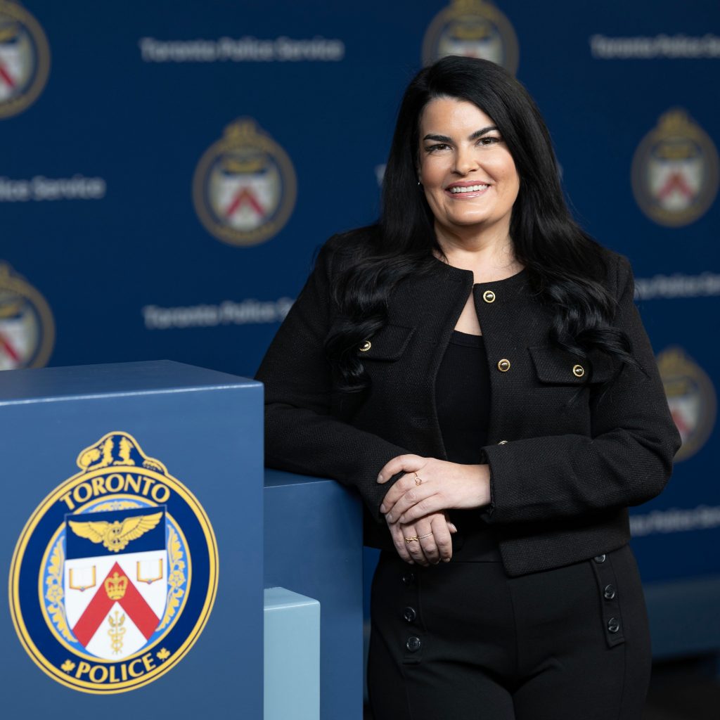 Person stands beside podium with Toronto Police Service emblem; blue backdrop behind features repeated Toronto Police Service logos throughout
