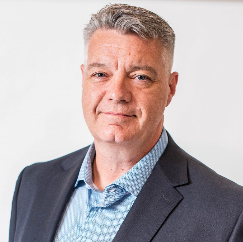Individual in light blue dress shirt and dark blazer with short gray hair, standing against plain white background