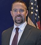 a man in a dark suit, white dress shirt, and red tie standing in front of an American flag. The background is a solid dark color.