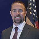 a man in a dark suit, white dress shirt, and red tie standing in front of an American flag. The background is a solid dark color.
