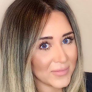 Person with straight, shoulder-length hair featuring dark roots and blonde highlights, wearing a dark top and a delicate necklace against a neutral background