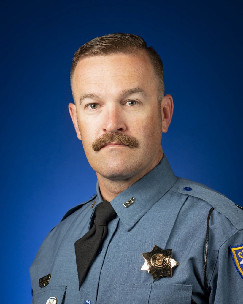 Person wearing a blue law enforcement uniform with black tie, silver collar pins, shoulder patch, and star-shaped badge stands against a solid blue backdrop