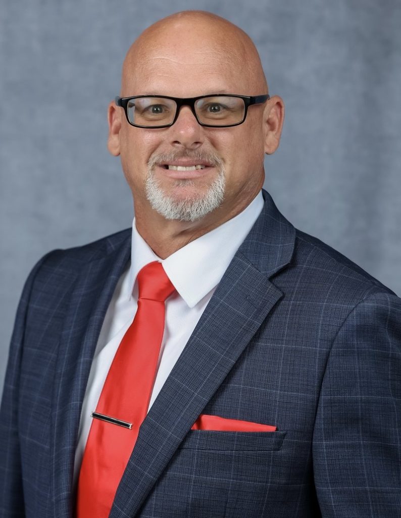 Studio portrait shows an individual in a gray checkered suit, white shirt, red tie with tie bar, and matching pocket square, posed against a neutral gray backdrop