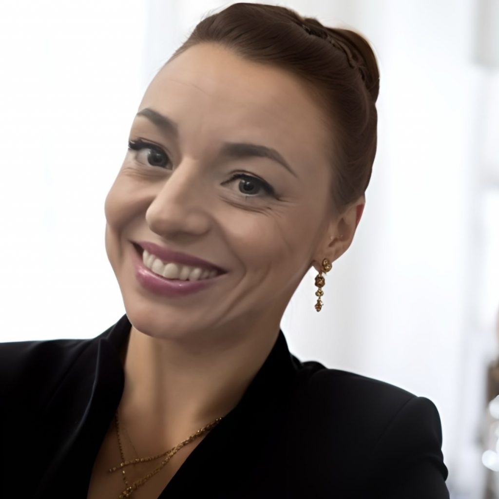 Person in black blazer with gold pendant necklace and dangling earrings stands before softly lit white curtains