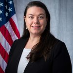 Person in black blazer over light gray top standing before a U.S. flag and gray backdrop in a formal portrait-style setting.