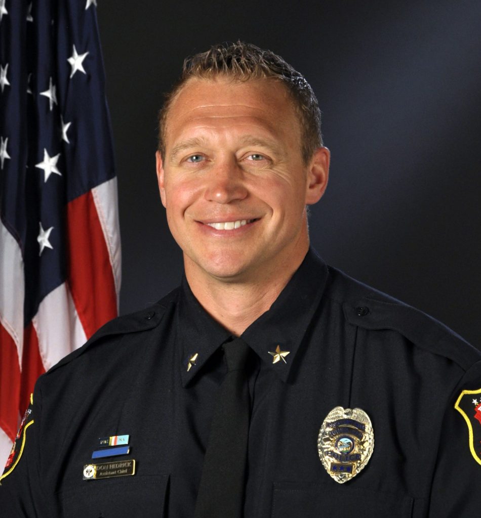 Person wearing a dark police dress uniform with silver badge, nameplate, and shoulder stars; U.S. flag in background and department patch on sleeve