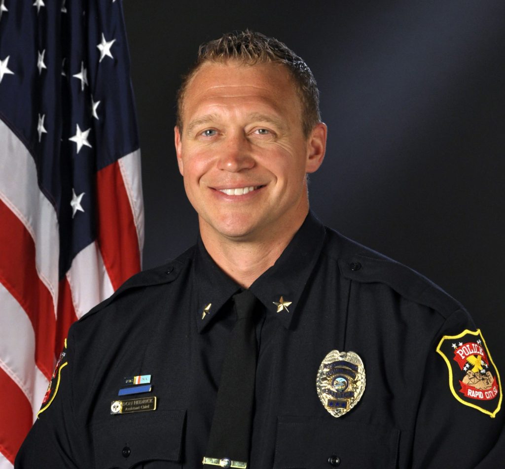 Police officer in dark uniform with gold badge, nameplate, and shoulder patch reading ‘Police,’ standing beside a U.S. flag against a black backdrop