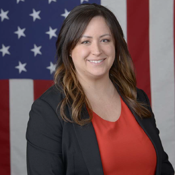 Individual in black blazer and red shirt stands before American flag backdrop with white stars on blue field and red-white stripes